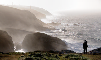 Looking out on cornish cliffs This landscape photograph, taken on a winter afternoon, captures the rugged cliffs of the Cornwall coast in the United Kingdom. The main subject of the image is a person looking out over the dramatic, mist-covered Cornish cliffs as waves crash against the rocky shoreline. In the background, several tall chimneys—reminiscent of Cornwall's historical mining heritage—rise above the cliffs, adding a distinctive feature to the scene. The photograph showcases the raw power of nature along this iconic coastline, with Gerald Weinbren’s composition emphasizing the interplay of light, sea spray, and rock formations.